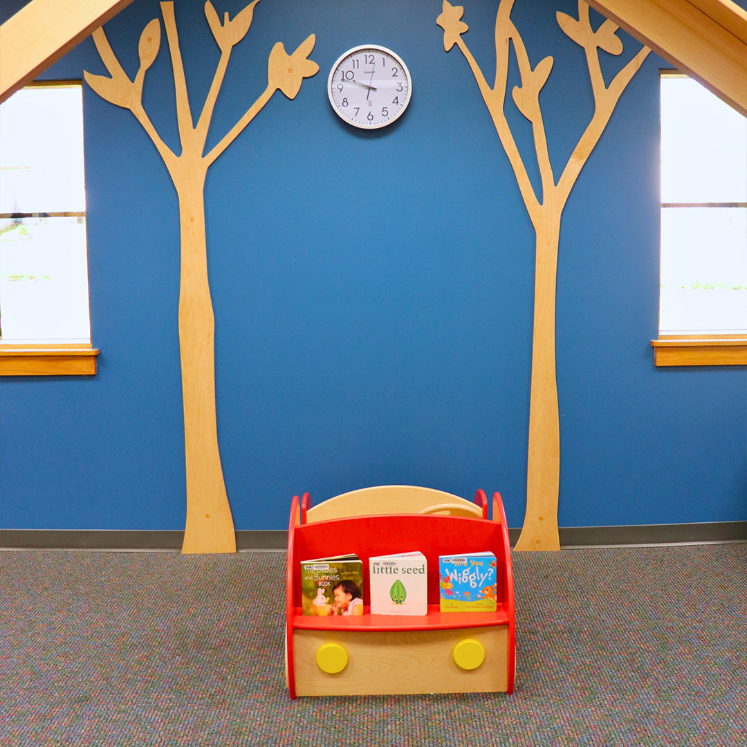 A small red climb-in car with a shelf for children’s books on the front sits in front of a blue wall between two wooden trees attached to the wall.
