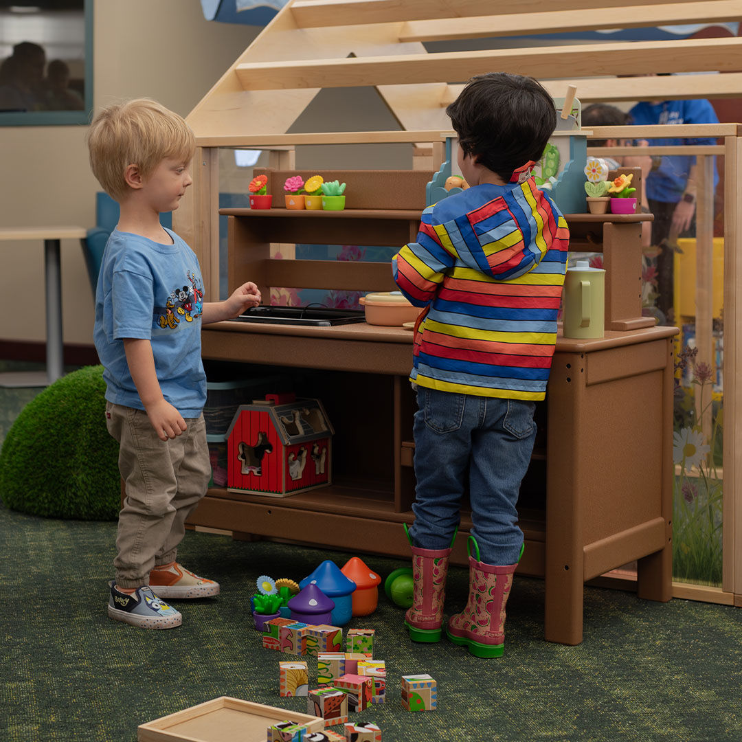 Two young children playing with a toy kitchen in the Storyscape area, with blocks and toy vegetables scattered on the floor.
