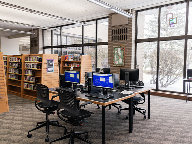 El interior de la biblioteca cuenta con grandes ventanales con vistas a un paisaje nevado, estaciones de ordenador con sillas en el centro y estanterías a su alrededor.