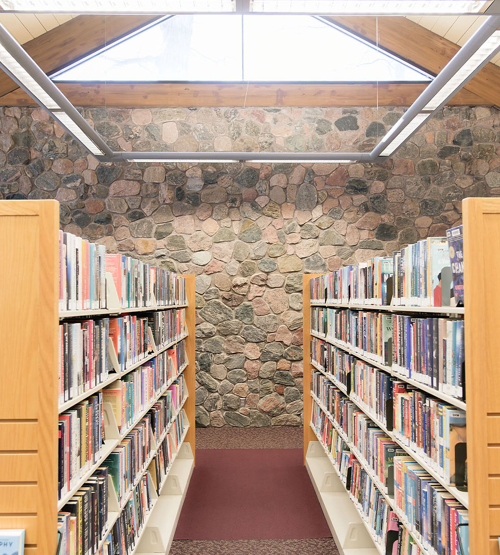 Un pasillo de biblioteca con estanterías de madera llenas de libros coloridos, una pared de piedra al final, una ventana triangular en la parte superior y lámparas colgantes rectangulares.