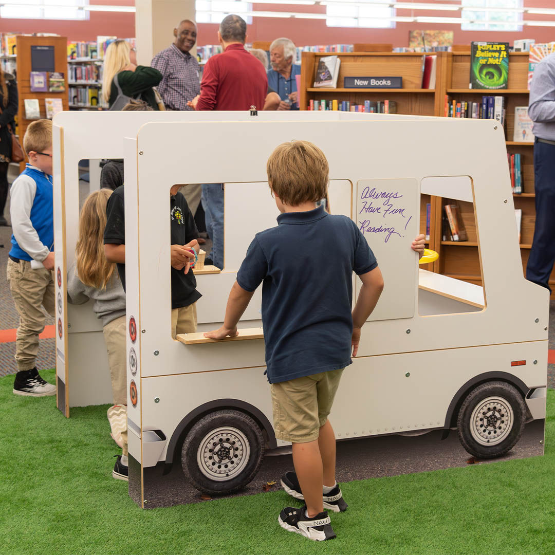 Children play inside a white wooden food truck in a library Storyscape area, with bookshelves and smiling adults visible in the background.