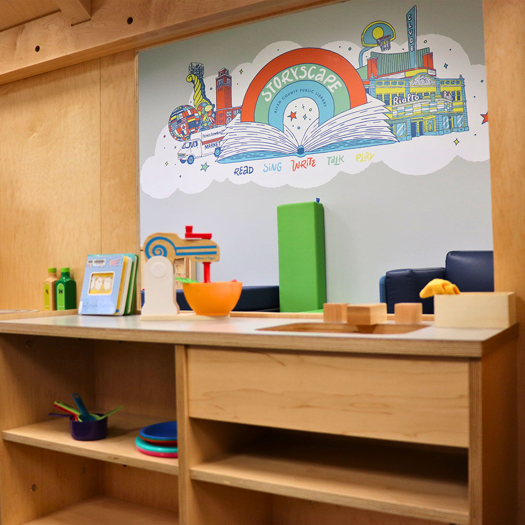 A Storyscape area featuring a Storyscape logo mural surrounded by playful illustrations. A wooden play kitchen in the foreground showcases toys and books.