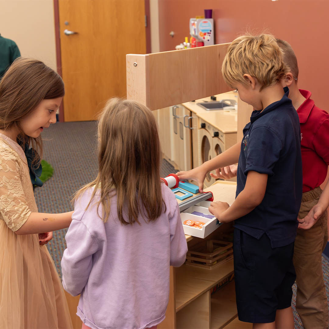 Children engaged in imaginative play at a pretend market stand, using a toy cash register, sharing, and smiling.