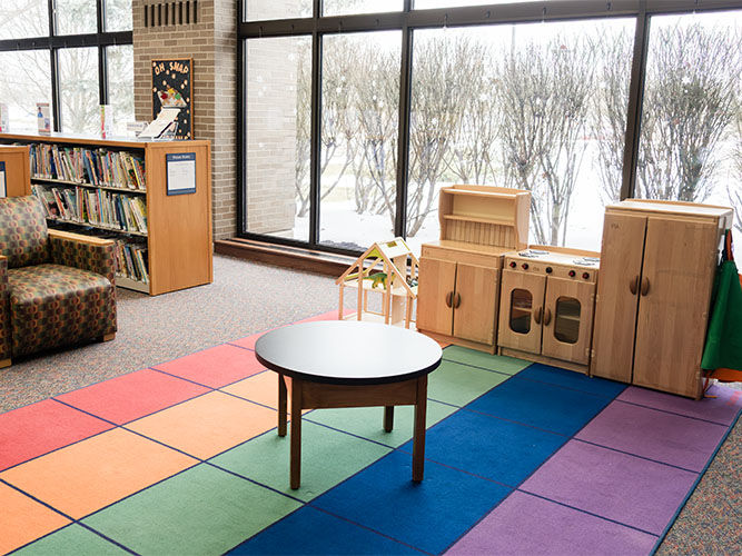 Zona de biblioteca infantil con alfombra a cuadros de arcoíris, mesa redonda, cocinita de juguete de madera y estanterías.