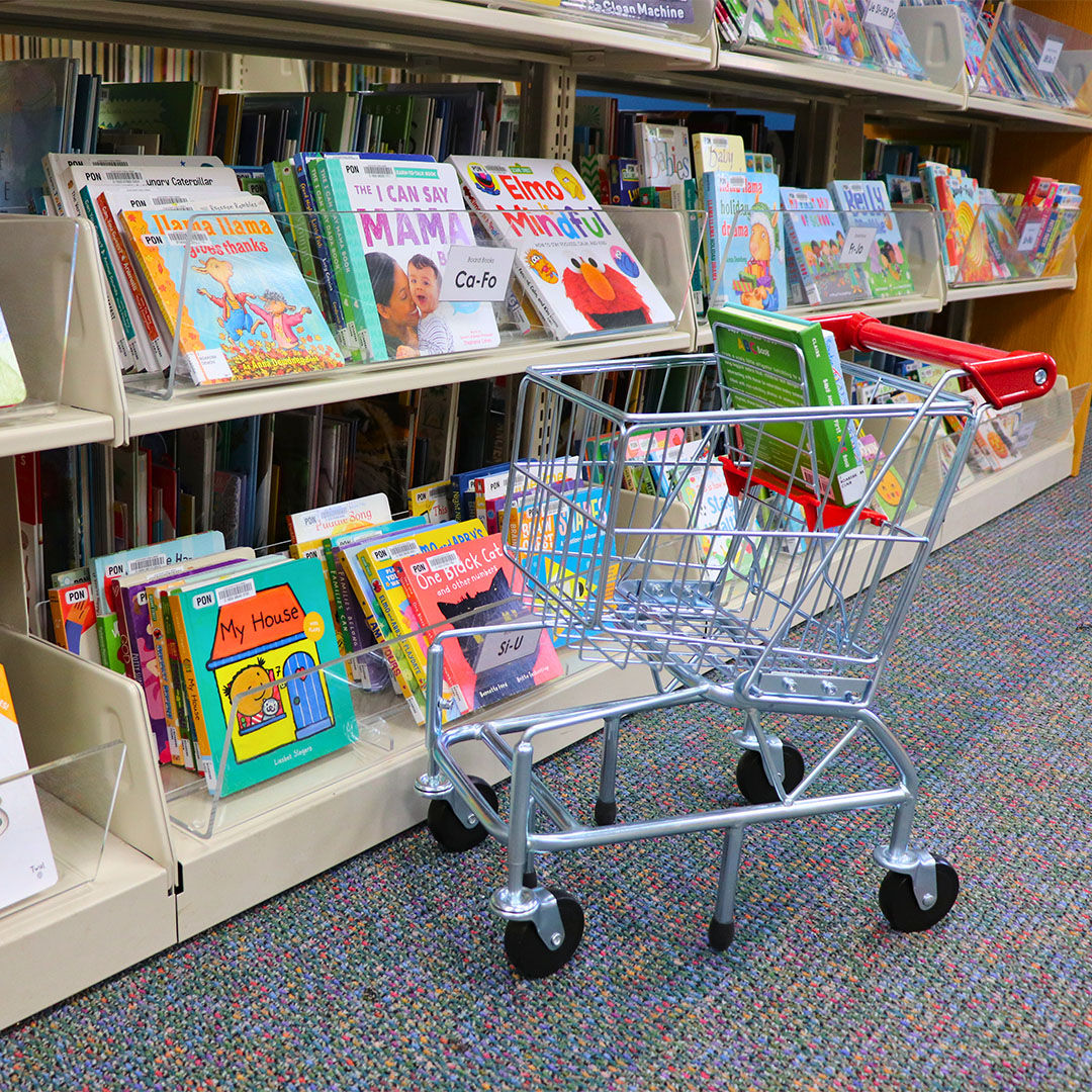 A child-sized shopping cart with a red handle sits by a library shelf filled with colorful children’s books.