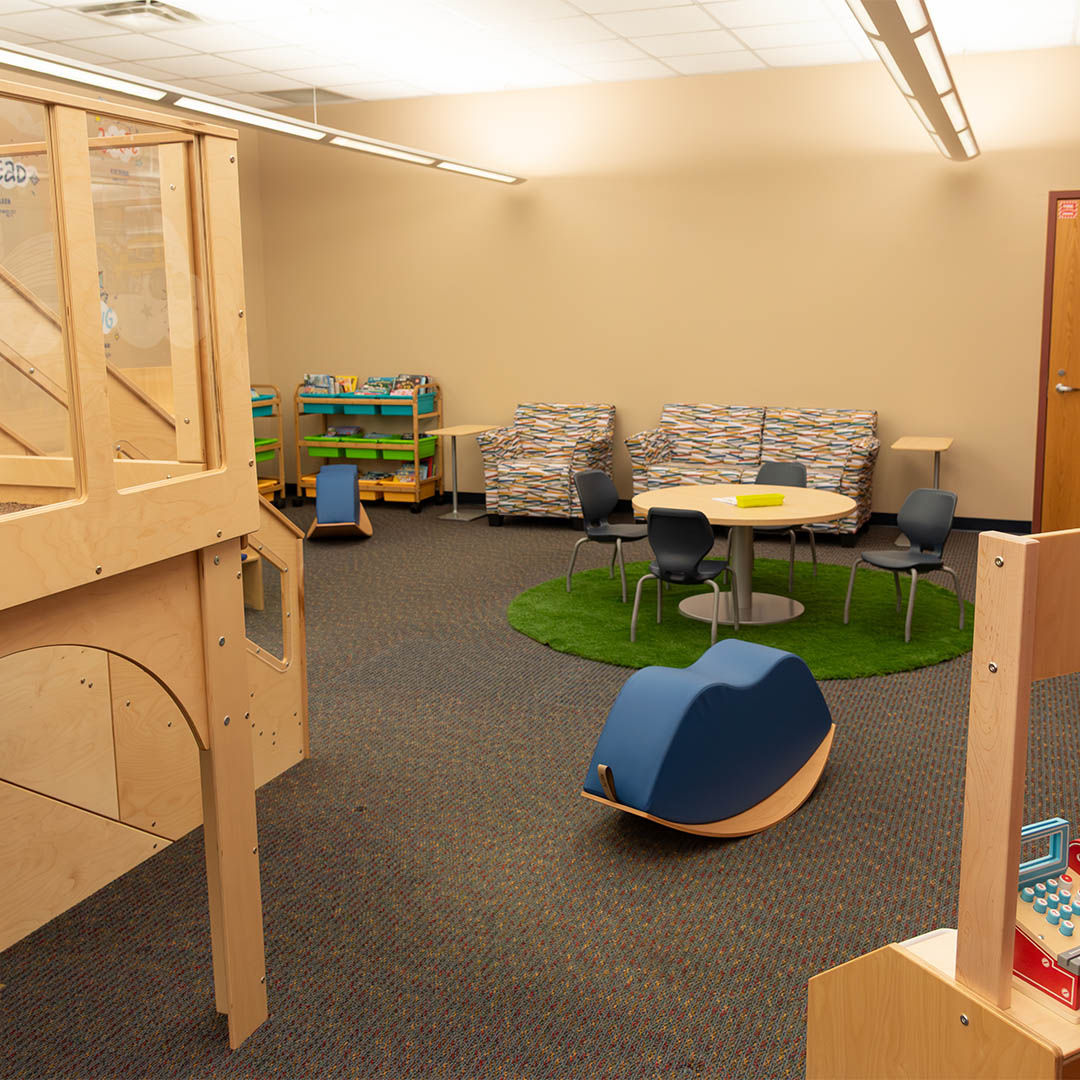 Overview of part of the Storyscape area with a wooden climbable play structure, colorful shelves, a round table with blue chairs on a green rug, a curved rocking seat for children, and cozy patterned couches.