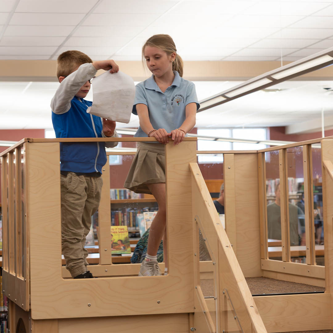 A young boy and girl stand on a wooden indoor play structure in a library. The boy examines a scavenger hunt sheet while the girl looks on curiously.