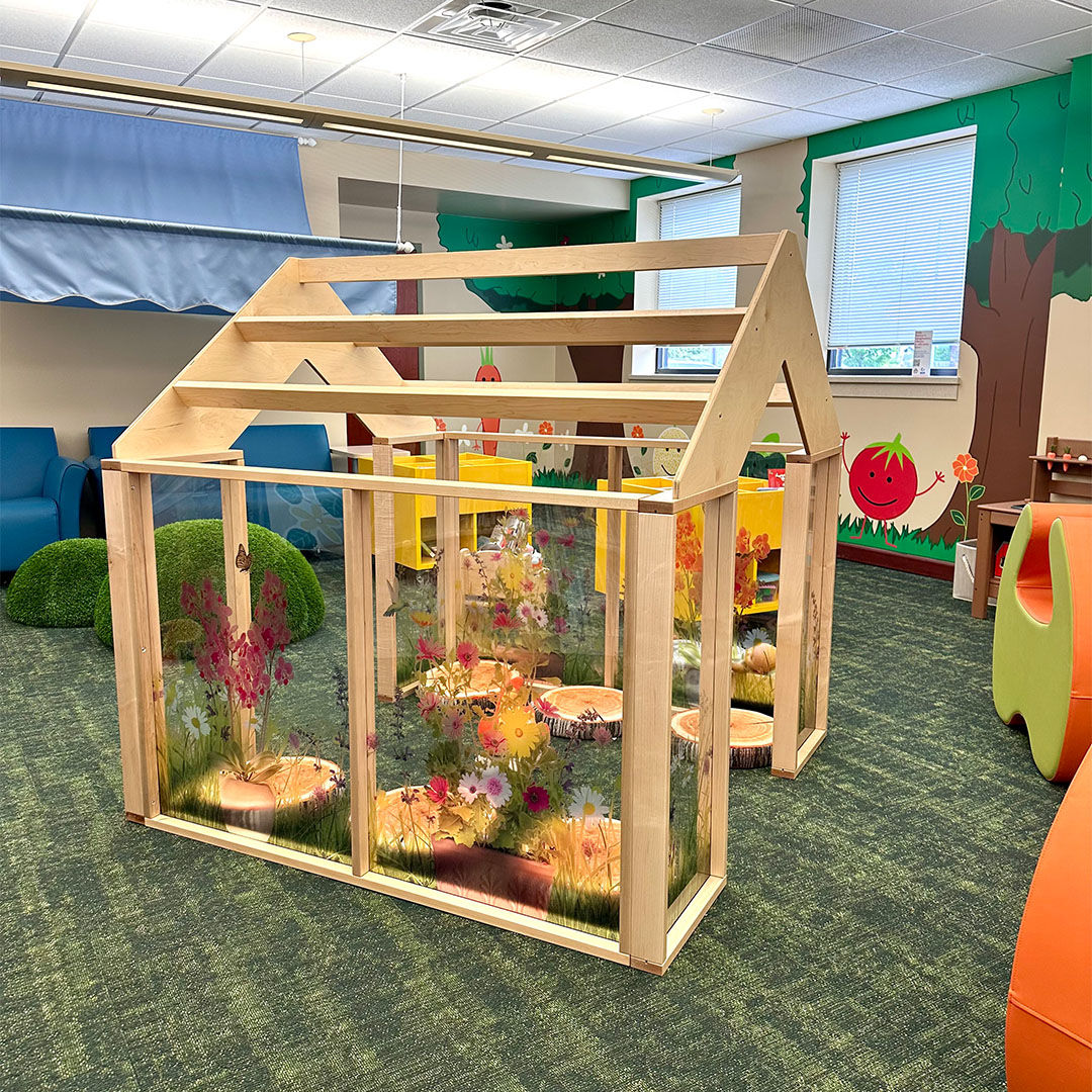 The Storyscape area featuring a wooden greenhouse with transparent panels decorated with colorful flowers, with faux tree stumps inside for children to use as soft seating.
