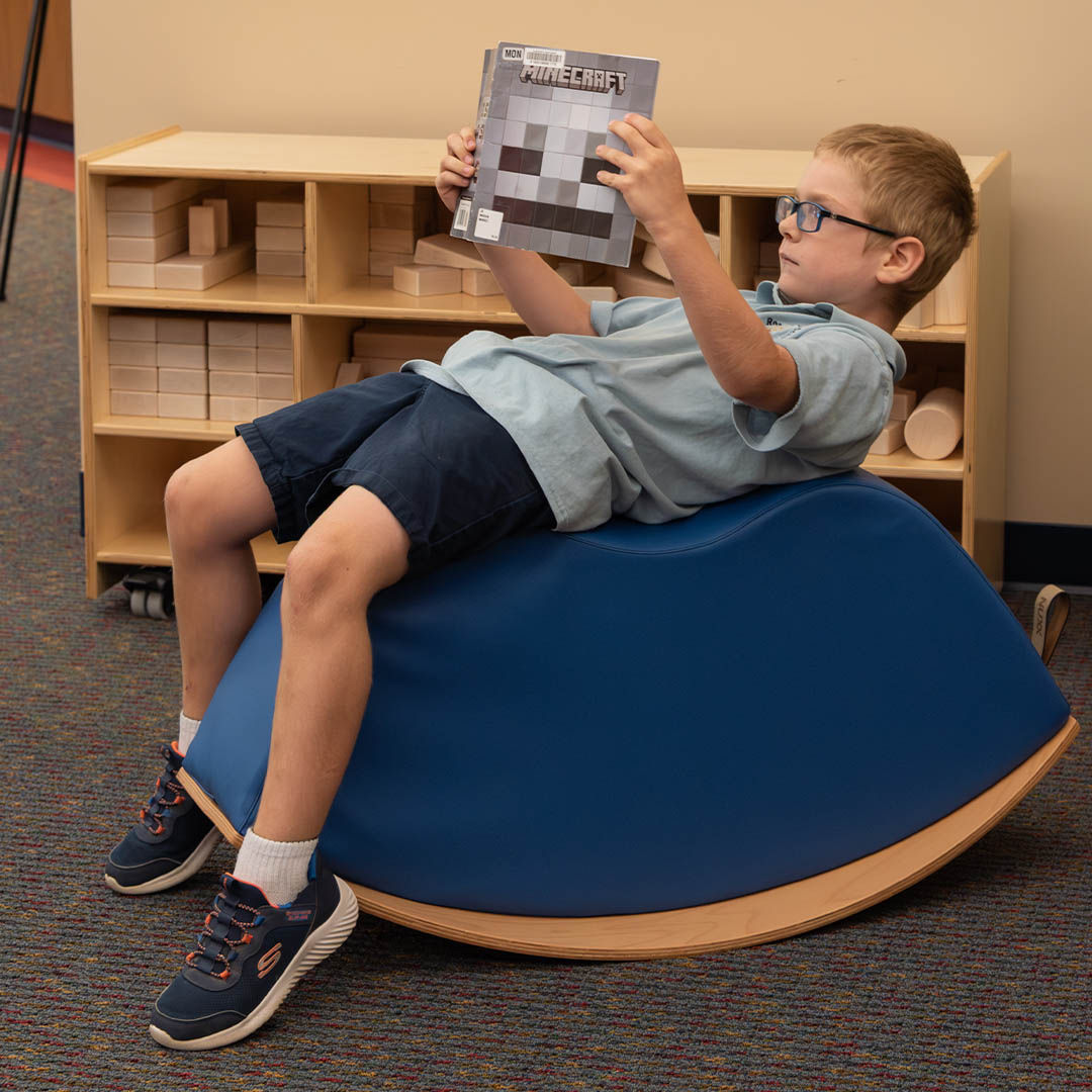 A child in glasses reclines on a blue curved chair while reading a Minecraft book. Wooden shelves with block toys are visible in the background.