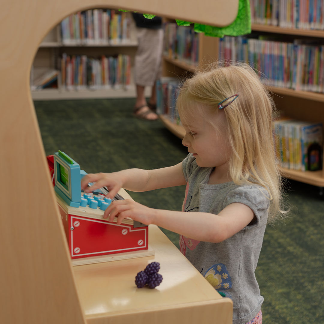 A young blonde-haired girl playing at a toy register in a library Storyscape section, with shelves of colorful books in the background.