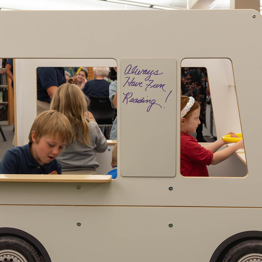 Children engaged in activities inside a playful white wooden food truck. A sign reads Always have fun reading.