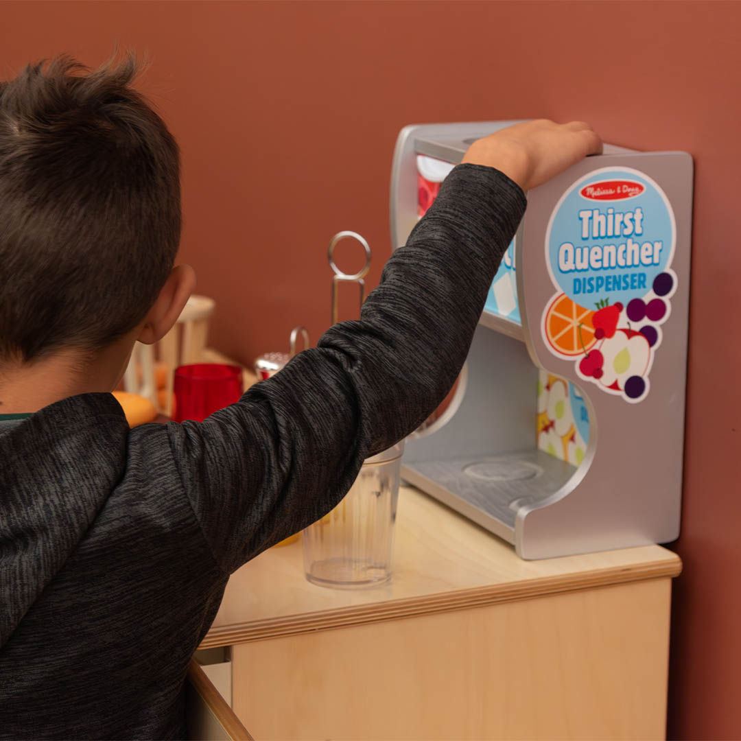 Young boy using a play juice dispenser labeled Thirst Quencher to fill a cup.