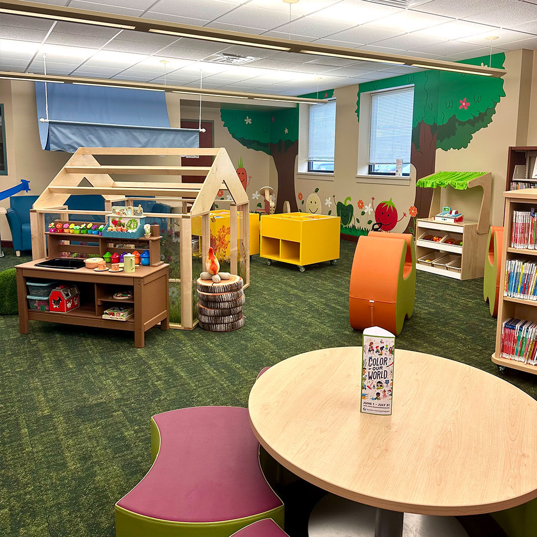 Colorful Storyscape area with a wooden greenhouse, toys, and books. Bright murals of trees and smiling fruits decorate the wall.